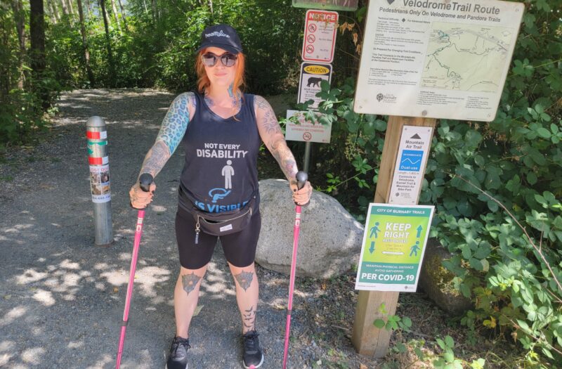 A photo of a woman, Eileen Davidson, standing in front of the entrance to a hike. She is holding hiking poles.