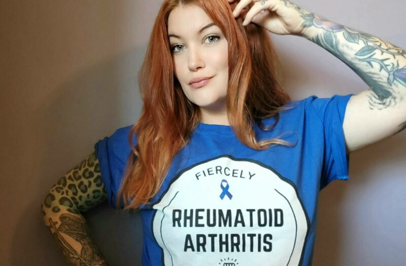 A photo of a woman, Eileen Davidson, modeling the "Fiercely Fighting Rheumatoid Arthritis" t-shirts, which will be available for purchase following the Tumbler and Tipsy by Michael Kuluva 2022 Collection event, airing on Tuesday, September 14, 20201 at 8 p.m. ET via YouTube. Eileen is wearing a blue shirt with a white circle outlined in black. Within the white circle are the words "Fiercely." Beneath that is a blue ribbon to symbolize arthritis awareness. Below the ribbon are the words "Rheumatoid Arthritis." Below those words are an image of a hand in a fist with lightning bolts. Below the hand is the worth "Fighting.